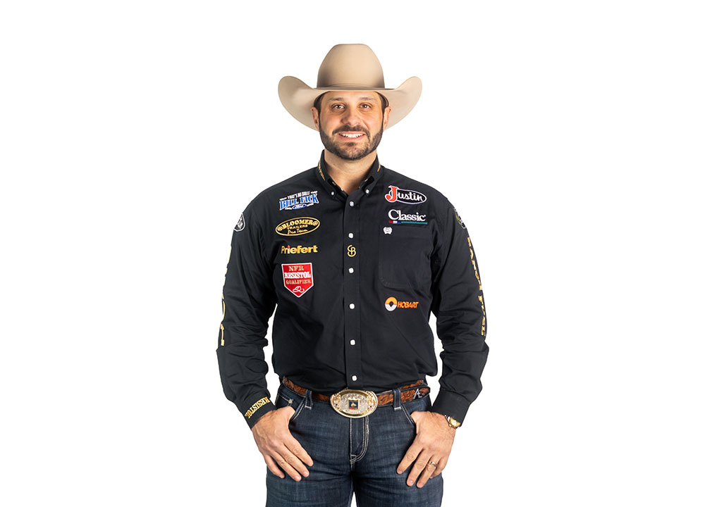 Anthony Lucia vistiendo una camisa negra y un sombrero de vaquero, sonriendo y posando frente a una pared blanca.